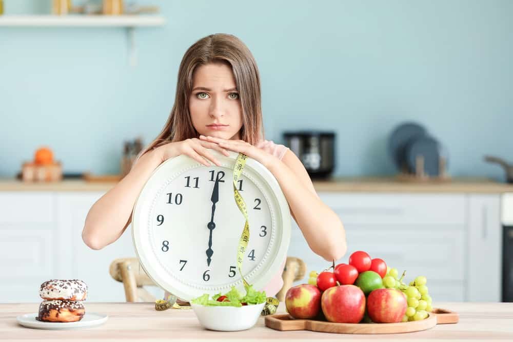 Sad woman with clock and food sitting in kitchen. Diet concept