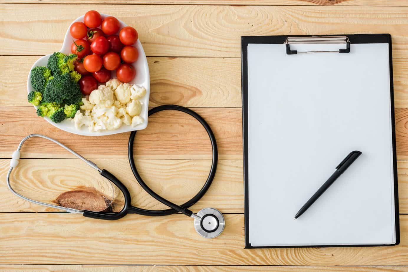 Top view of blank clipboard with pen near stethoscope and tasty vegetables on heart-shape plate