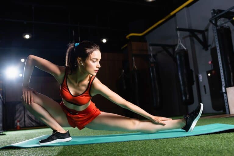 Sportswoman in top and shorts stretching legs on fitness mat in sports center