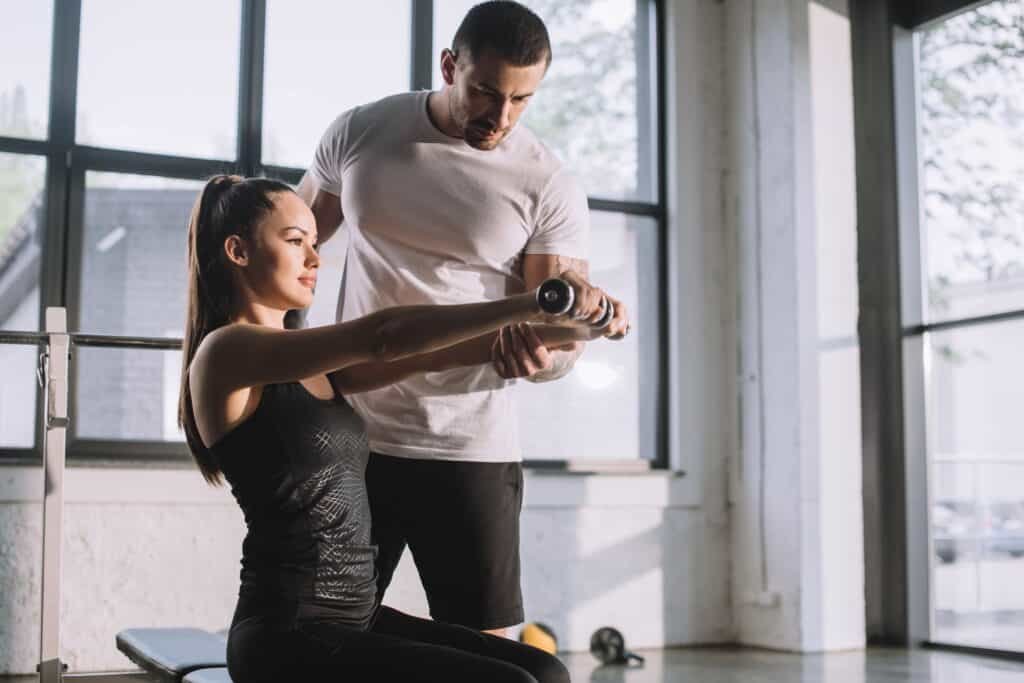 Why You Should Hire A Personal Trainer 2 Male personal trainer helping sportswoman to do exercises with dumbbells