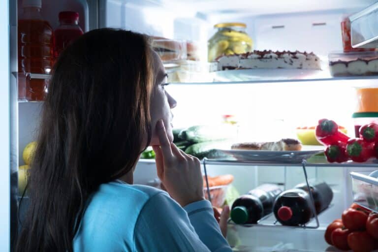 Close-up Of A Thoughtful Young Woman Looking for food in an open refrigerator