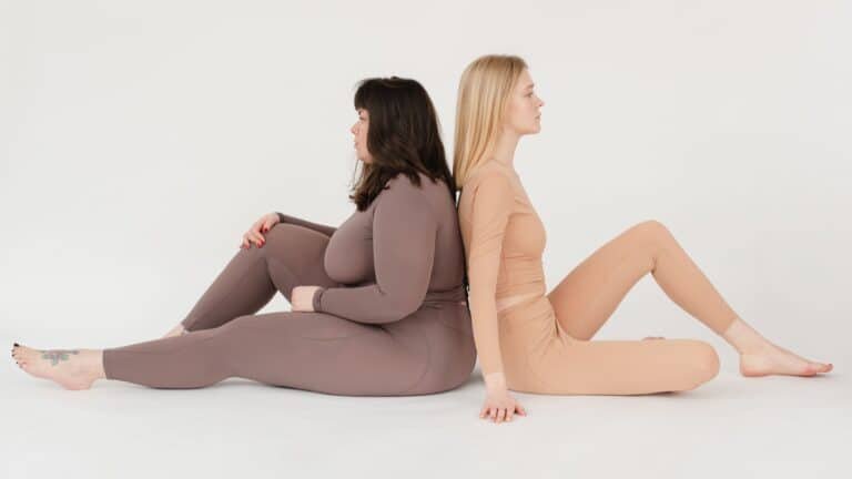 Young slim and obese woman sitting back to back in white studio